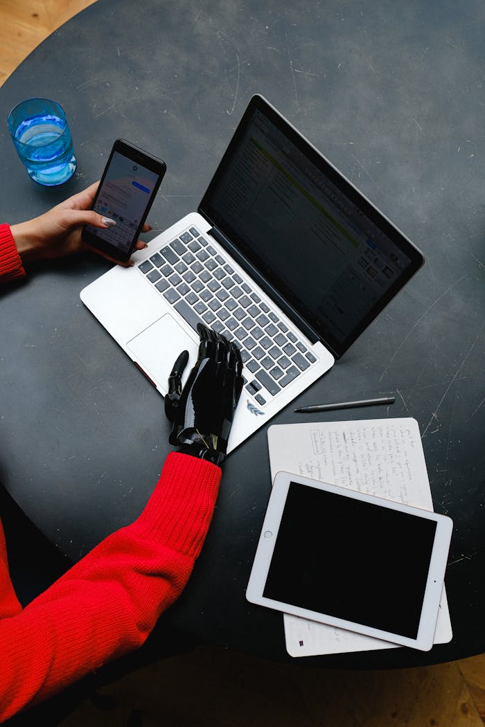 services-01 A prosthetic hand uses a laptop on a workspace with gadgets and notes around.